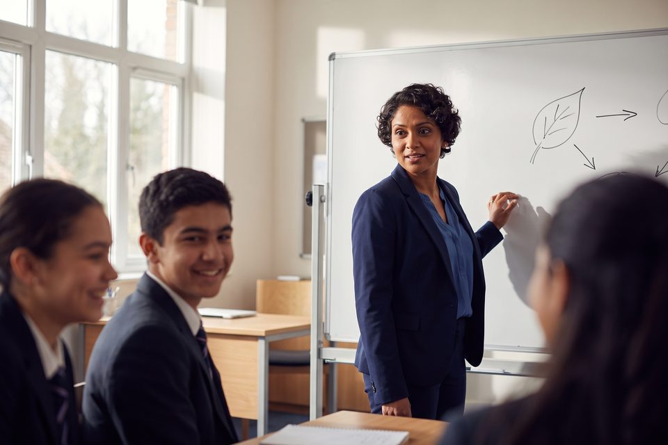 A teacher at a green board with a chalk sketch of a leaf turns to ask her class a question