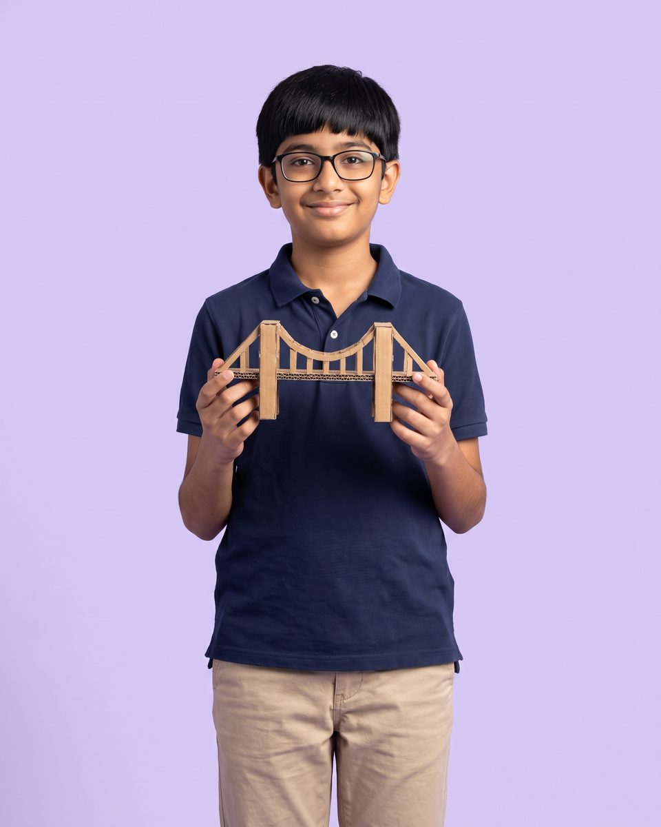 A 13-year-old boy in glasses proudly holds a model bridge
