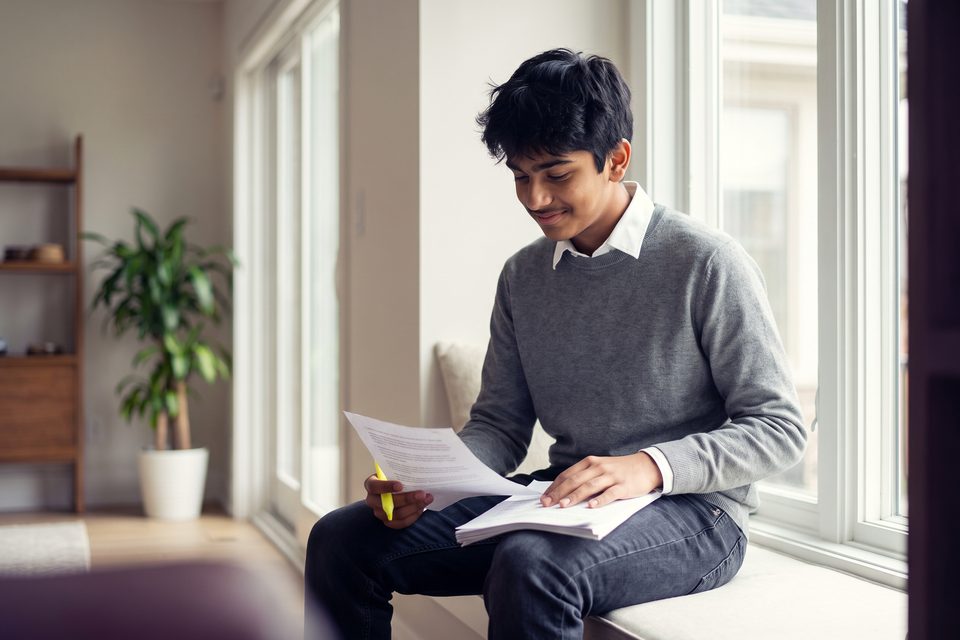 A teenage boy reads his notes with a highlighter on a sunny window seat
