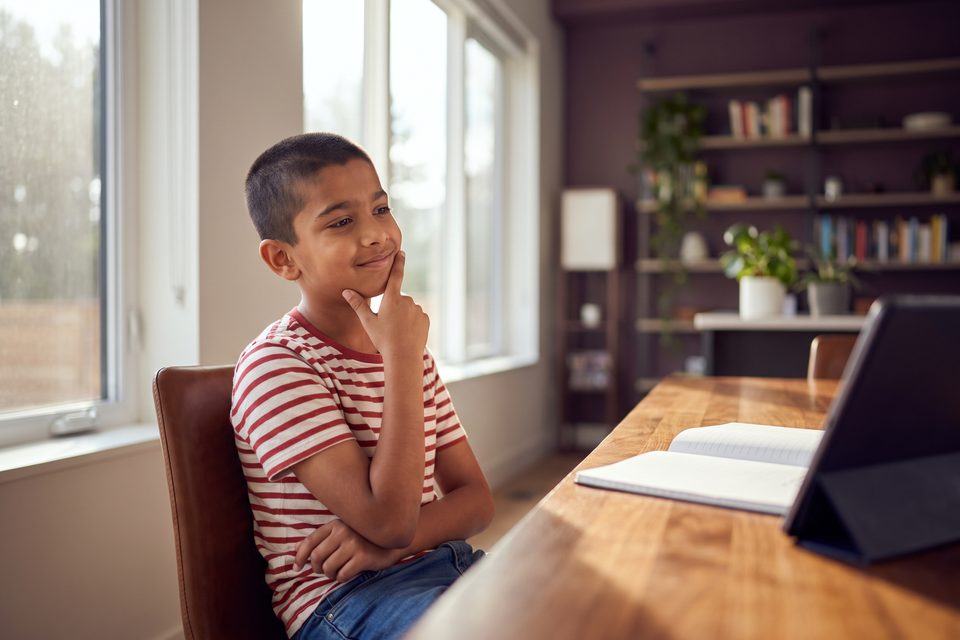 A boy thinks about a question at a bright desk, finger on his chin
