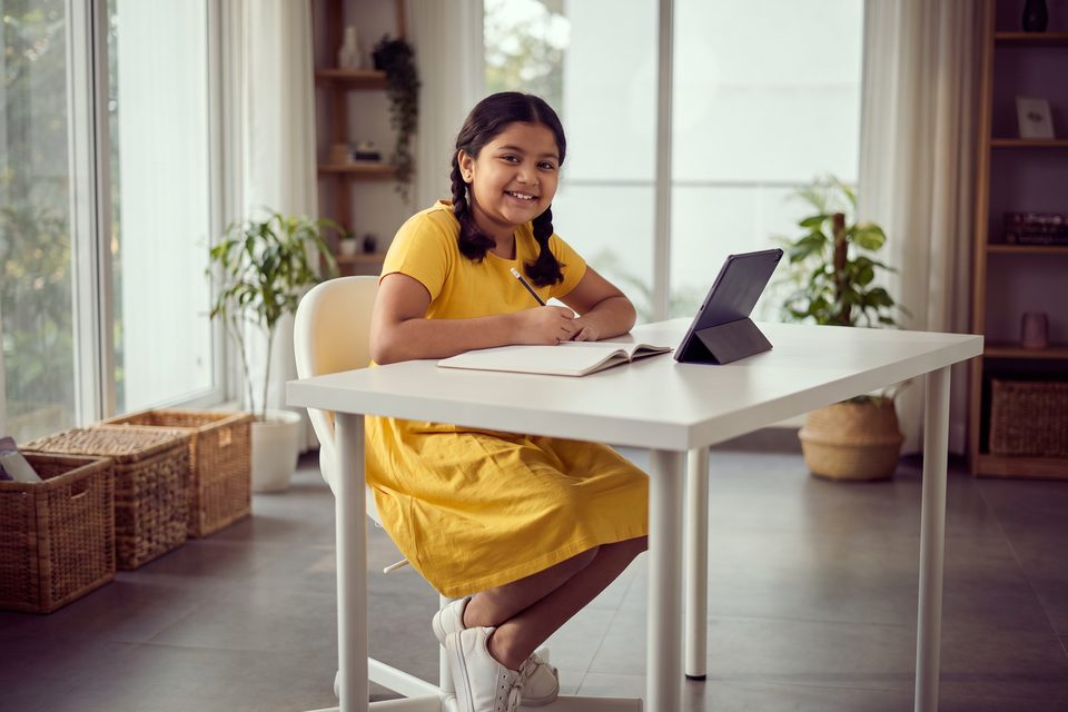 A smiling girl starts a lesson at a bright study desk with a tablet and notebook