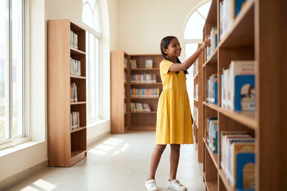 A smiling girl reaches for a book in a bright modern school library