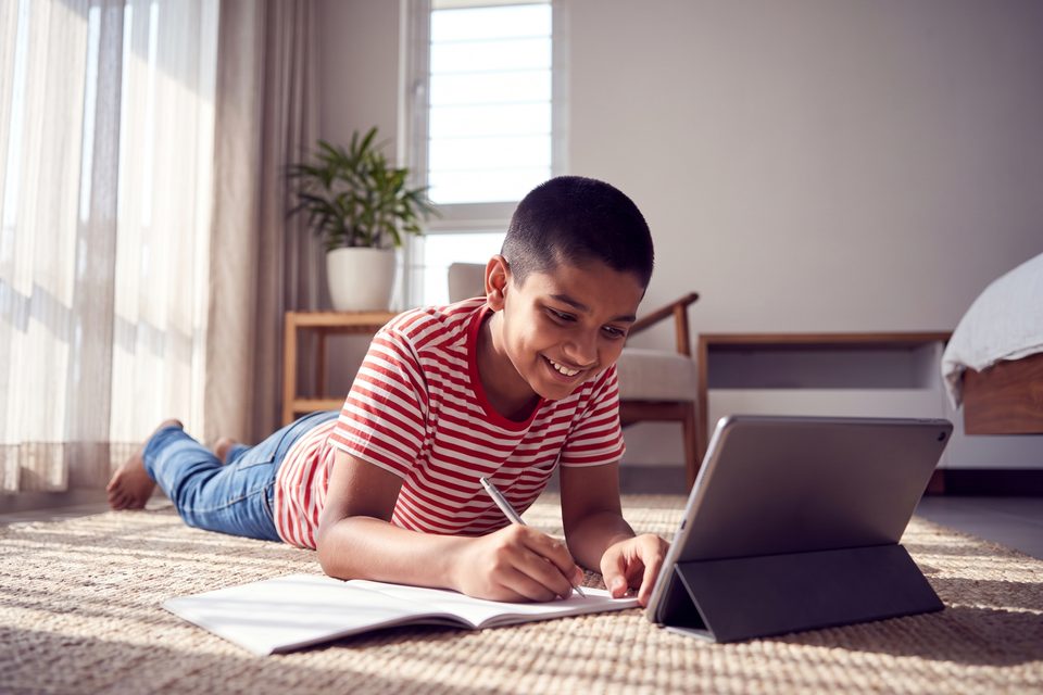 A boy lies on a bright bedroom rug learning on a tablet, smiling